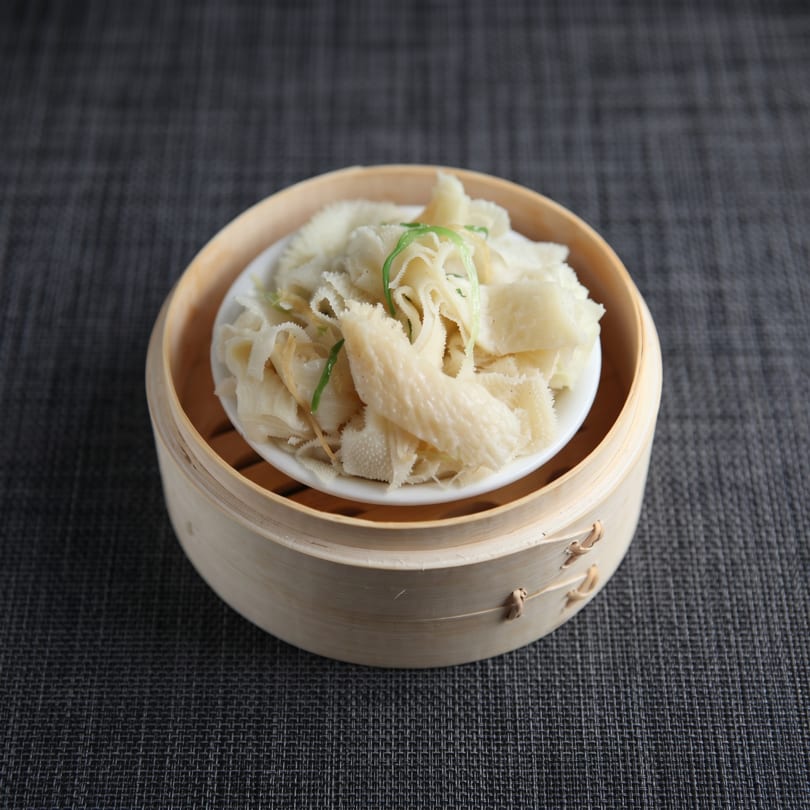 Steamed tripe with green onions in a bamboo dim sum basket on a dark grey tablecloth.