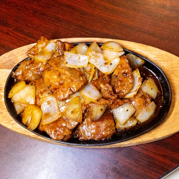 Sizzling beef and onions in a dark sauce, served on a black cast iron platter with a wooden base.