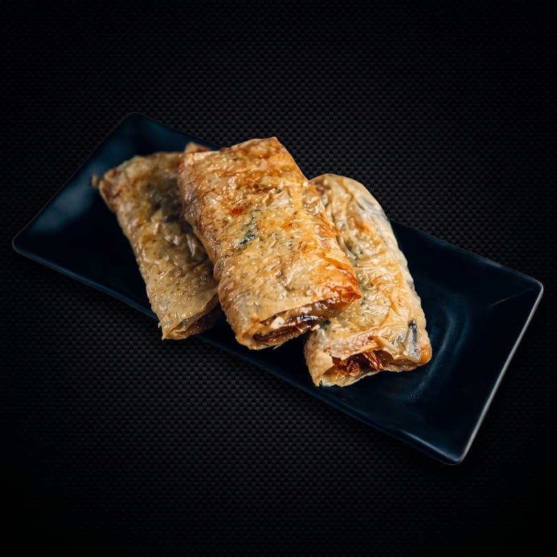 Three crispy, golden-brown spring rolls filled with vegetables, served on a dark rectangular plate.