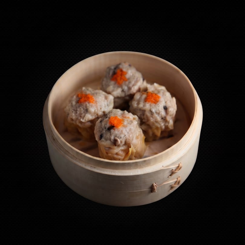 Four steamed dim sum dumplings with orange roe topping in a bamboo steamer basket.