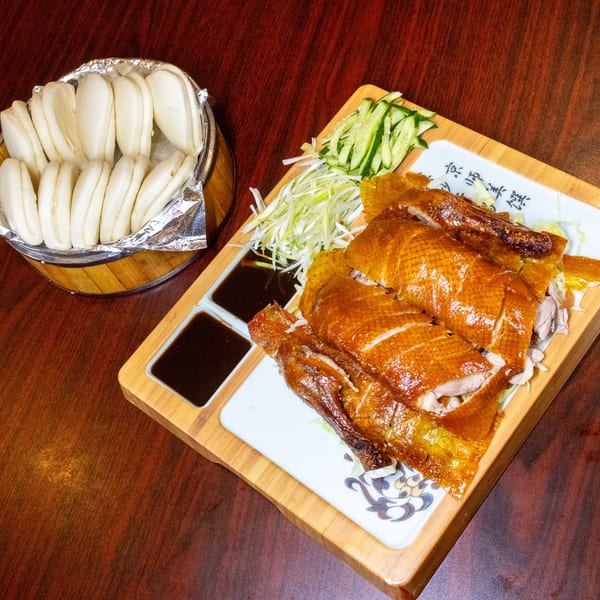 Peking duck with crispy skin, sliced cucumber, scallions, sauces, and steamed buns on a wooden table.