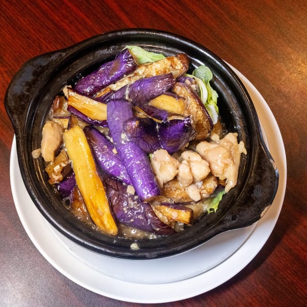 Clay pot dish with purple and yellow eggplant, chicken, and green lettuce on a wooden table.