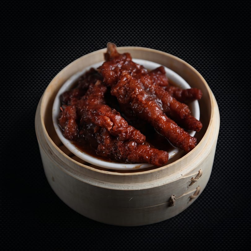Braised chicken feet in a rich, dark sauce, served in a white bowl within a bamboo steamer.