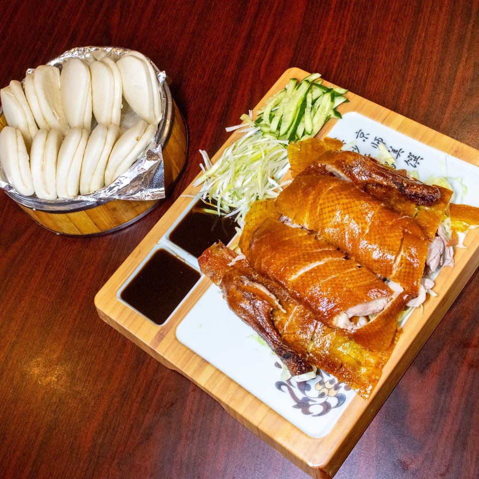 Peking duck with crispy skin, sliced cucumber, scallions, and steamed buns on a wooden table.