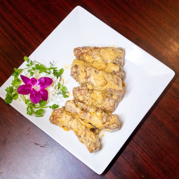 Crispy pork ribs coated in a creamy yellow sauce, garnished with a purple orchid and cilantro.