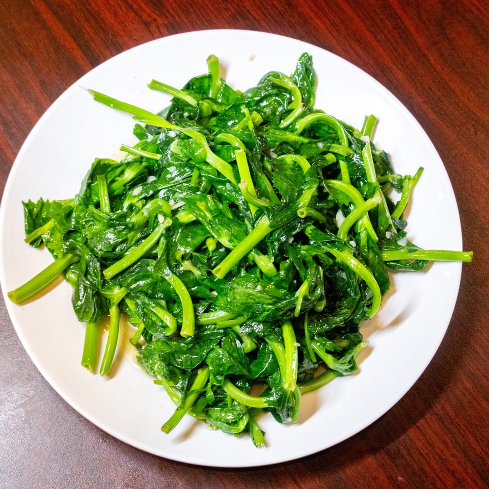 A white plate filled with vibrant green, sautéed leafy vegetables, possibly spinach or pea shoots, on a wooden table.